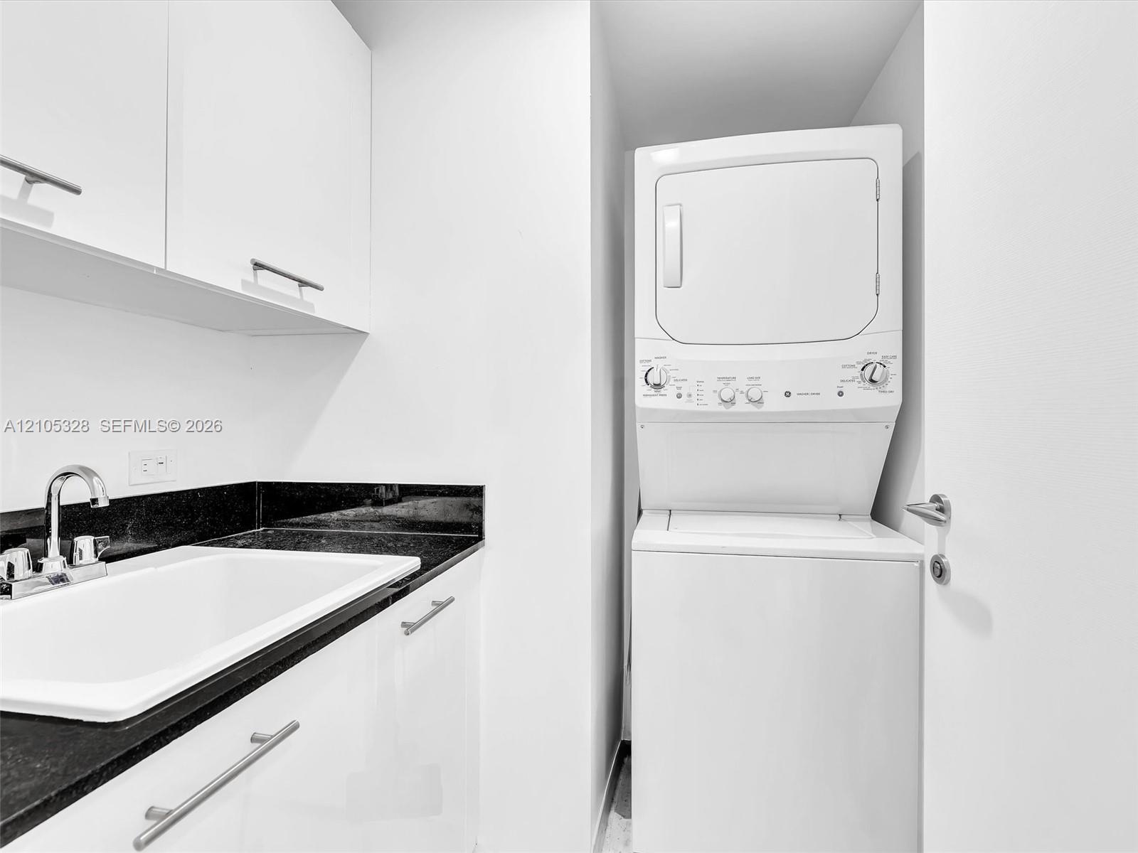 Stacked washer and dryer unit, white cabinetry with silver-tone pulls, black countertop, and an undermount sink with a chrome faucet