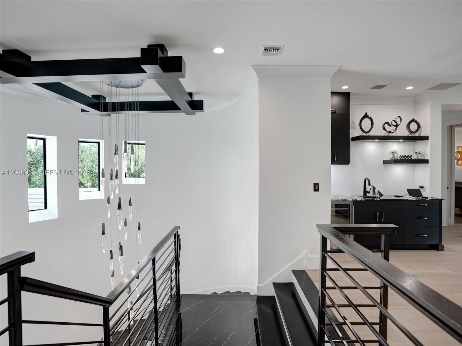 Black metal staircase railing with a multi-pendant glass chandelier, ceiling-mounted geometric accent structure, and a built-in wet bar featuring dark