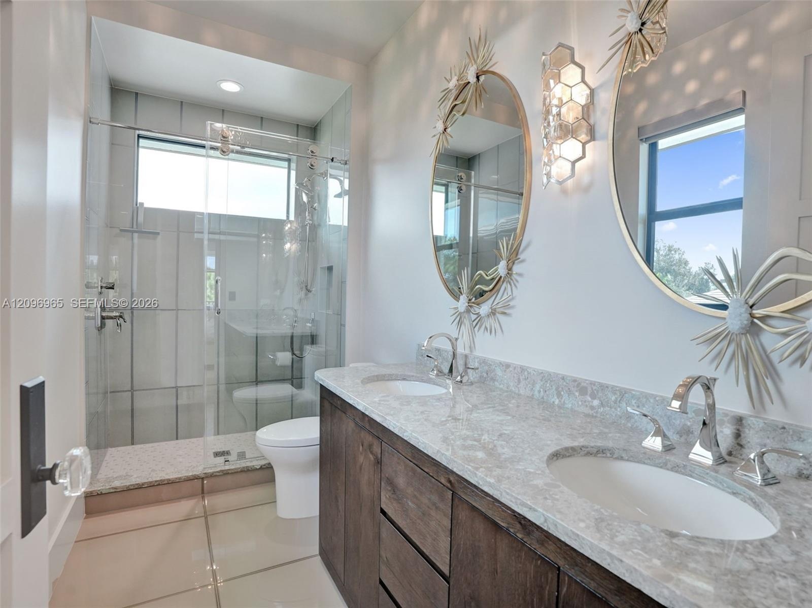 Dual-sink vanity with stone-finish countertop and wood-finish cabinetry, paired with a glass-enclosed shower featuring tile surrounds and wall-mounted