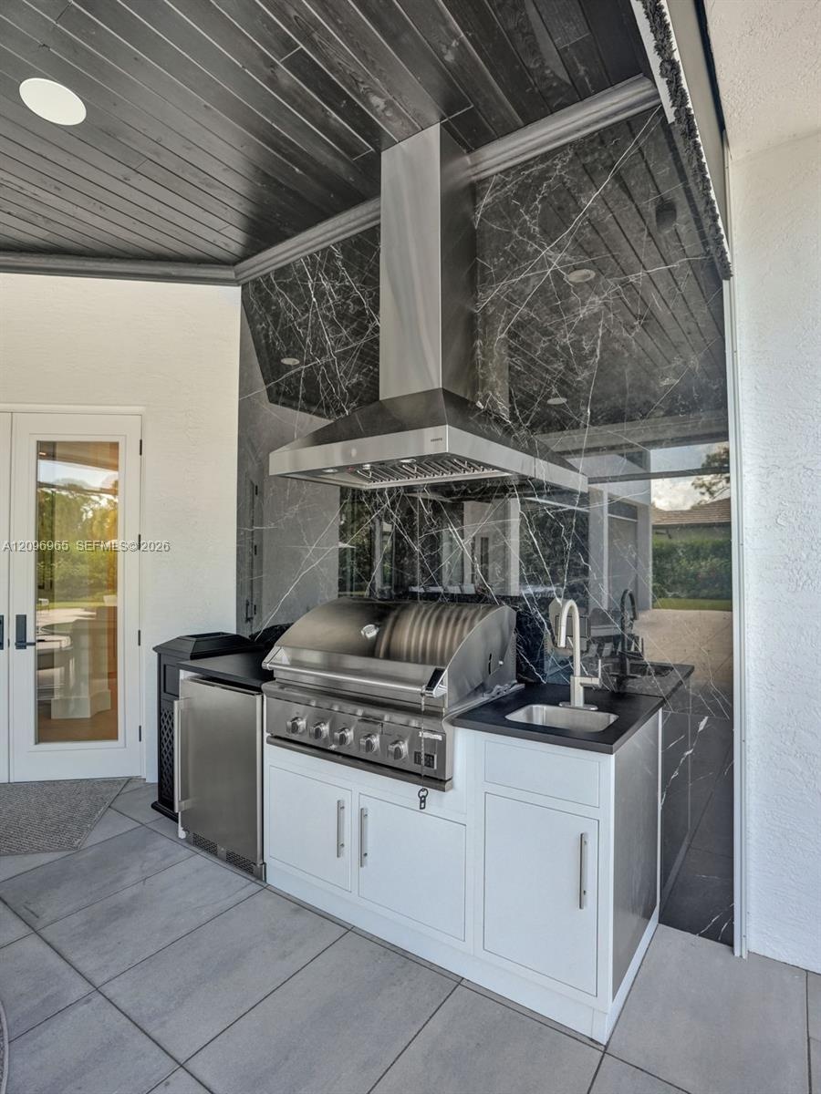 Outdoor kitchen featuring a stainless steel grill, under-counter refrigeration, prep sink, dark stone backsplash, and white cabinetry