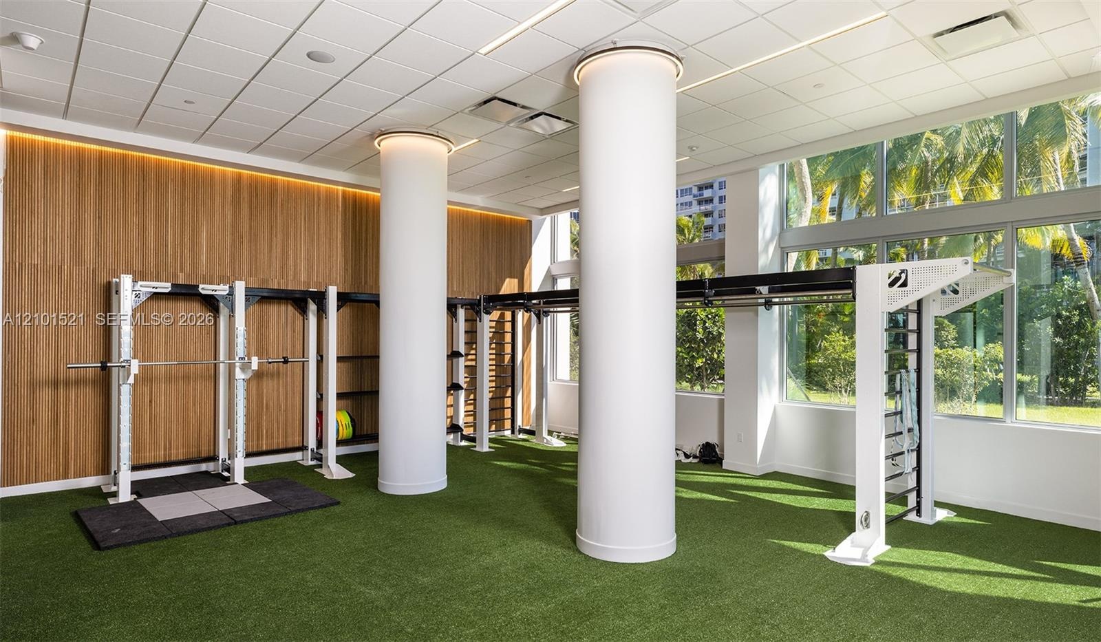 Fitness space featuring floor-to-ceiling windows, wood-paneled walls, synthetic turf flooring, recessed ceiling lighting, and structural cylindrical c