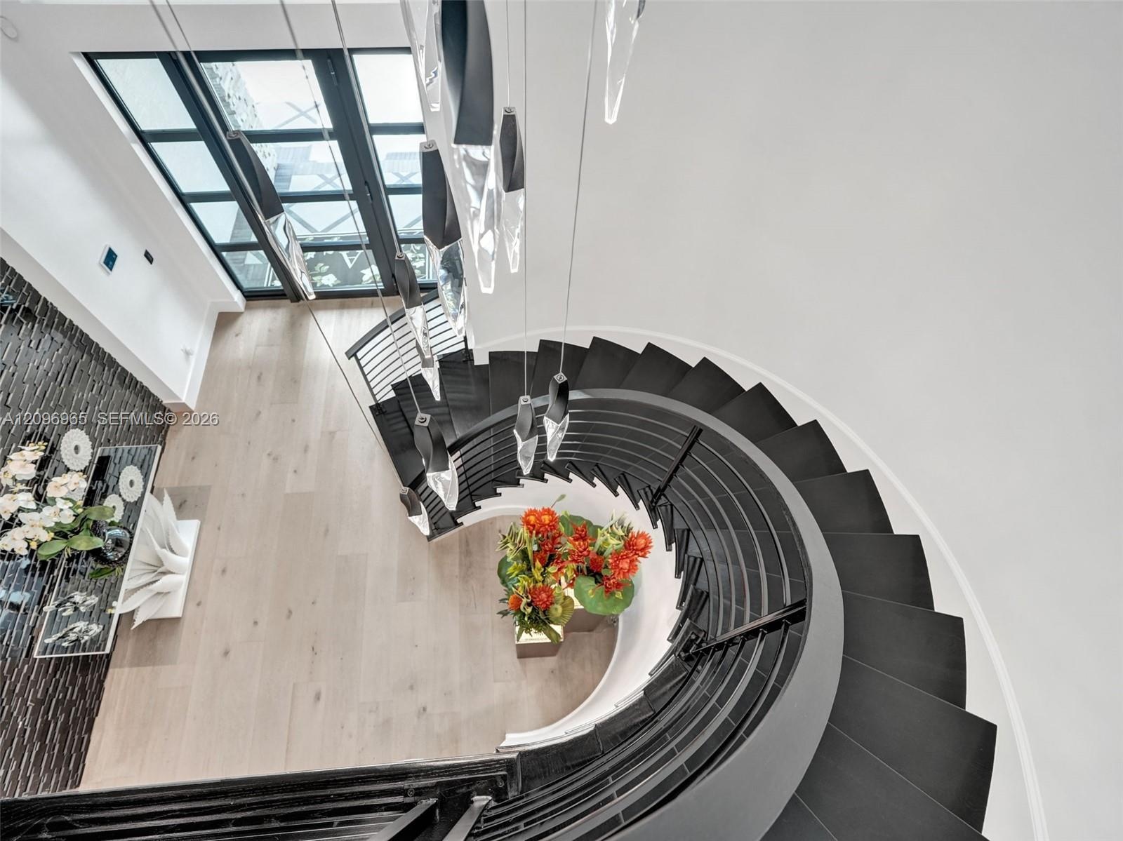Curved staircase featuring dark treads, black metal railing, floor-to-ceiling glass-paneled door, modern multi-pendant light fixture, and wood-finish 