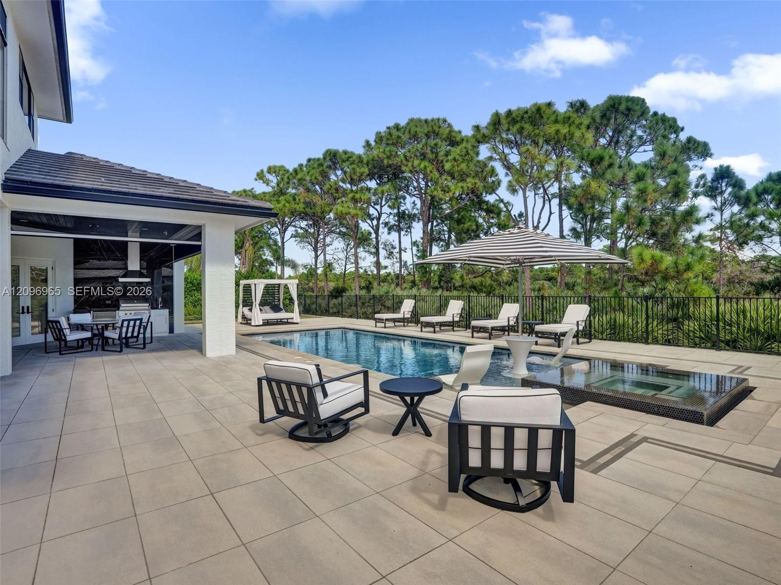 Expansive paved patio with a covered outdoor kitchen, rectangular swimming pool, raised spa, and black metal perimeter fencing