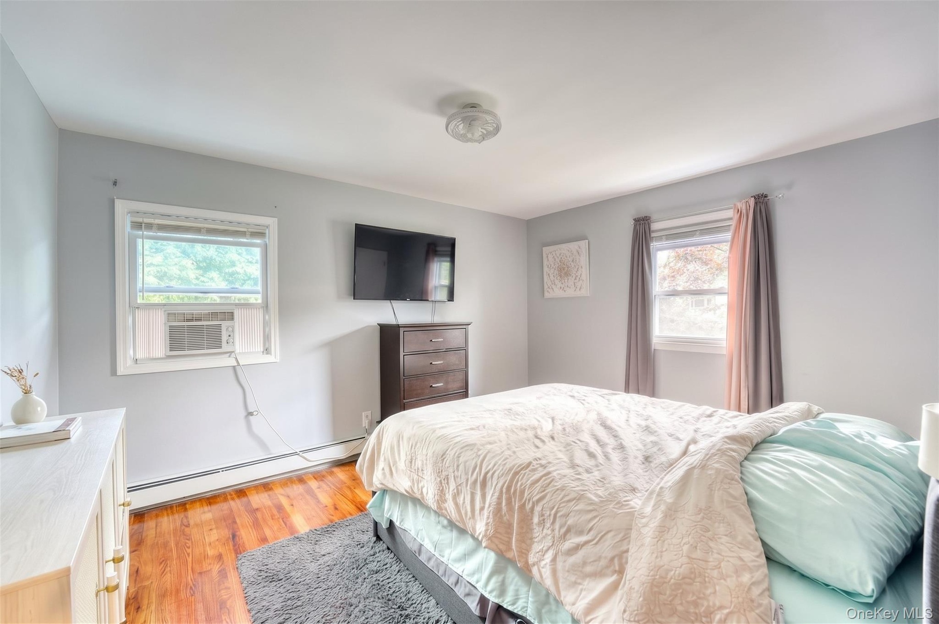 Bright and comfortable bedroom featuring hardwood floors, soft gray walls, and large windows that fill the space with natural light. The room includes