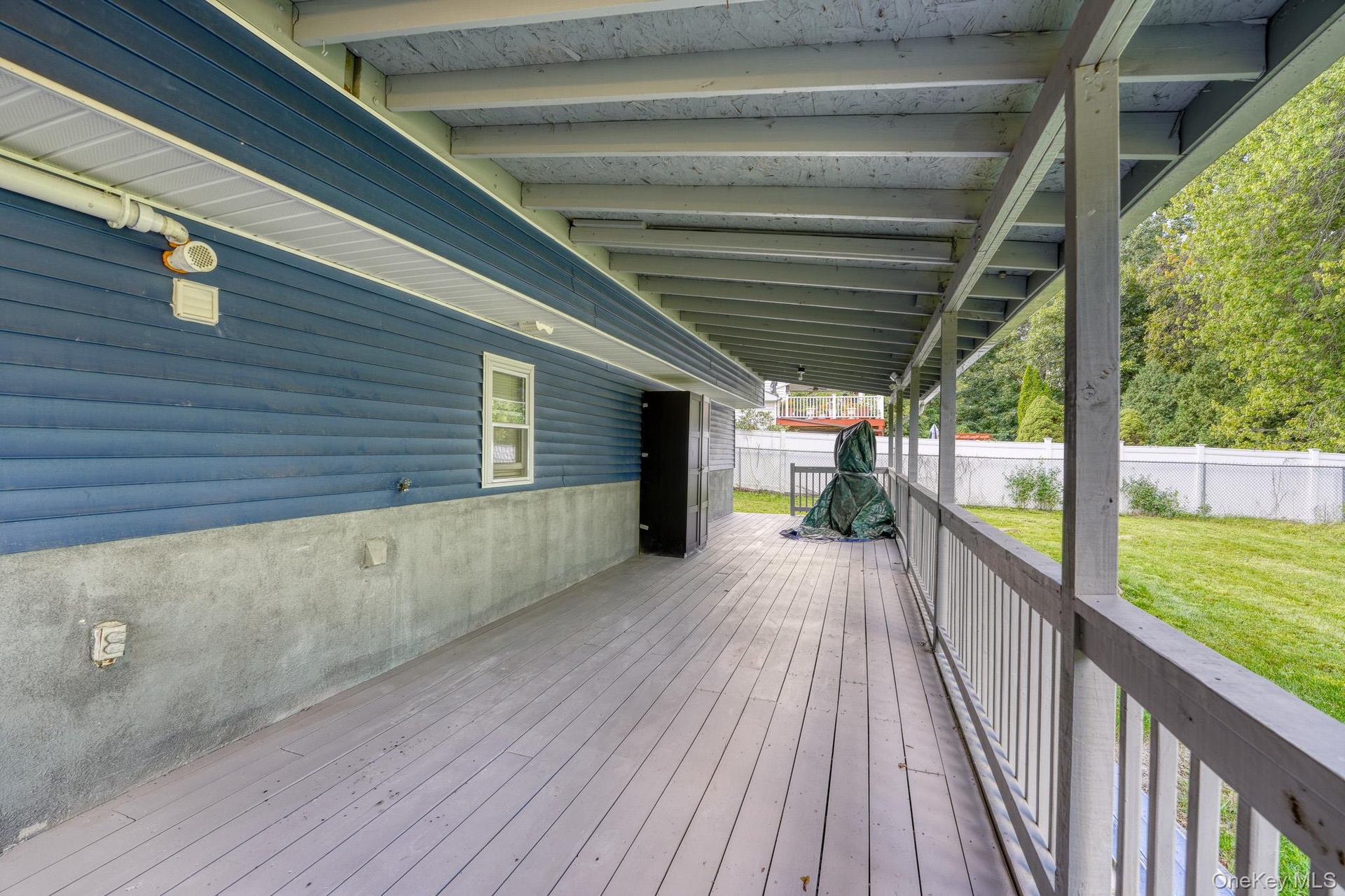 Expansive covered back porch with light gray decking and sturdy railing, offering a shaded retreat overlooking the backyard. The blue siding and expos