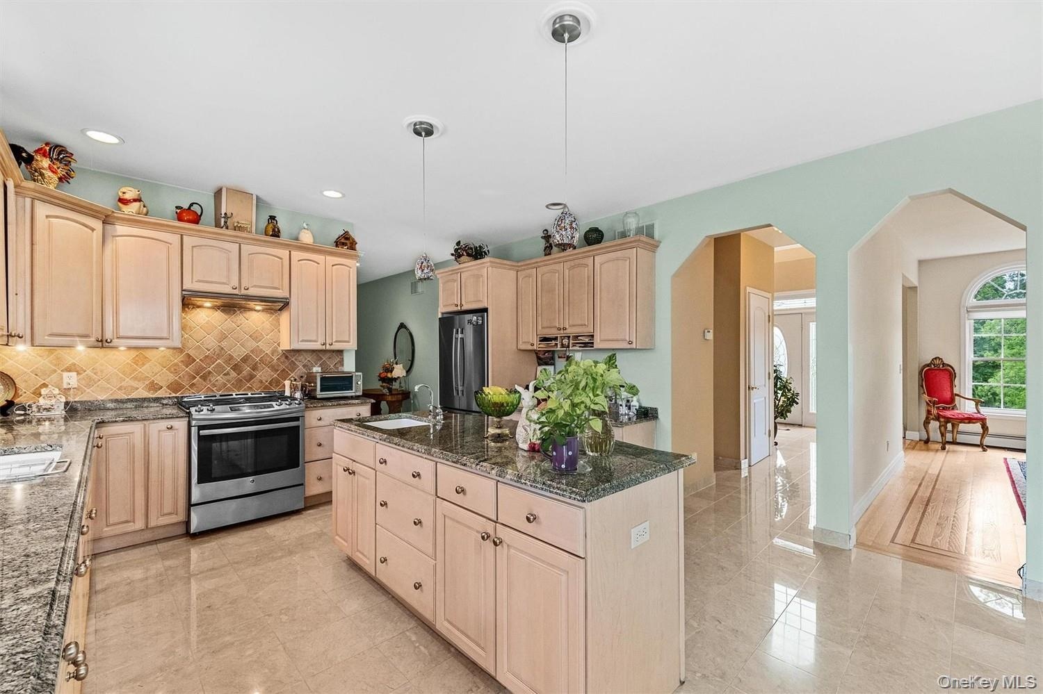 Kitchen featuring arched walkways, light wood finish cabinets, stainless steel appliances, dark stone countertops, and decorative light fixtures