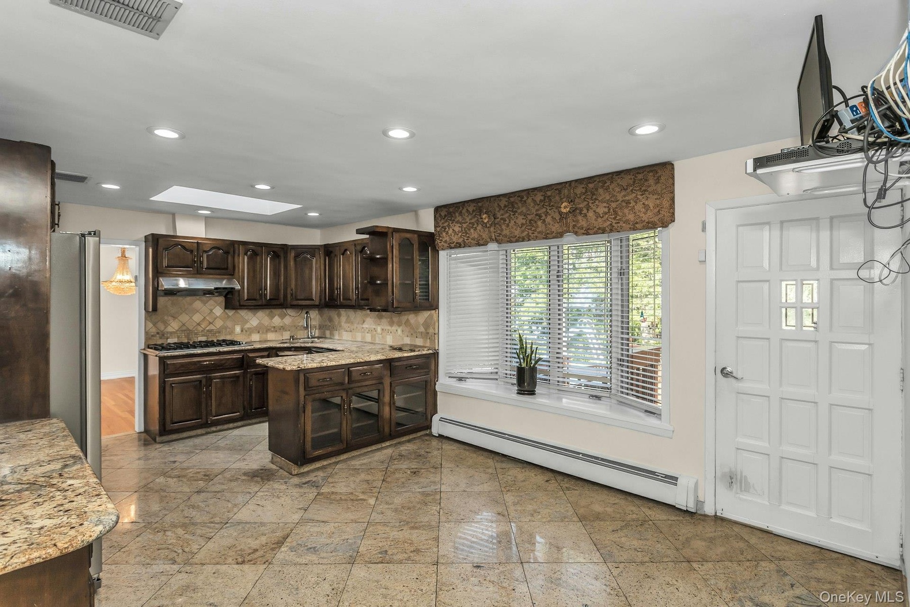 Kitchen featuring glass insert cabinets, dark brown cabinets, baseboard heating, light stone countertops, and decorative backsplash