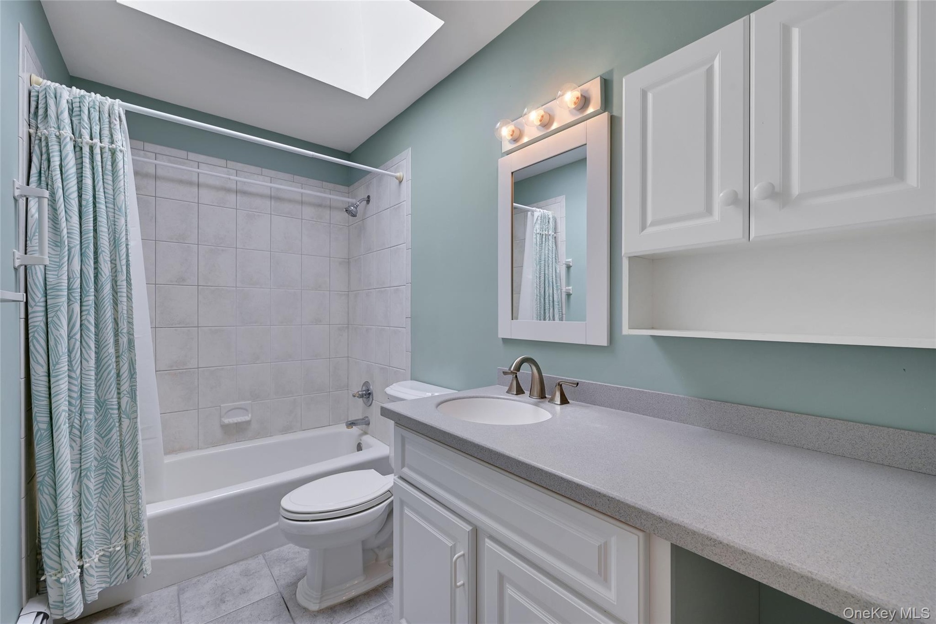 Main Hall Bathroom featuring a skylight, white tile shower surround, integrated vanity sink, and white cabinetry with brushed nickel hardware