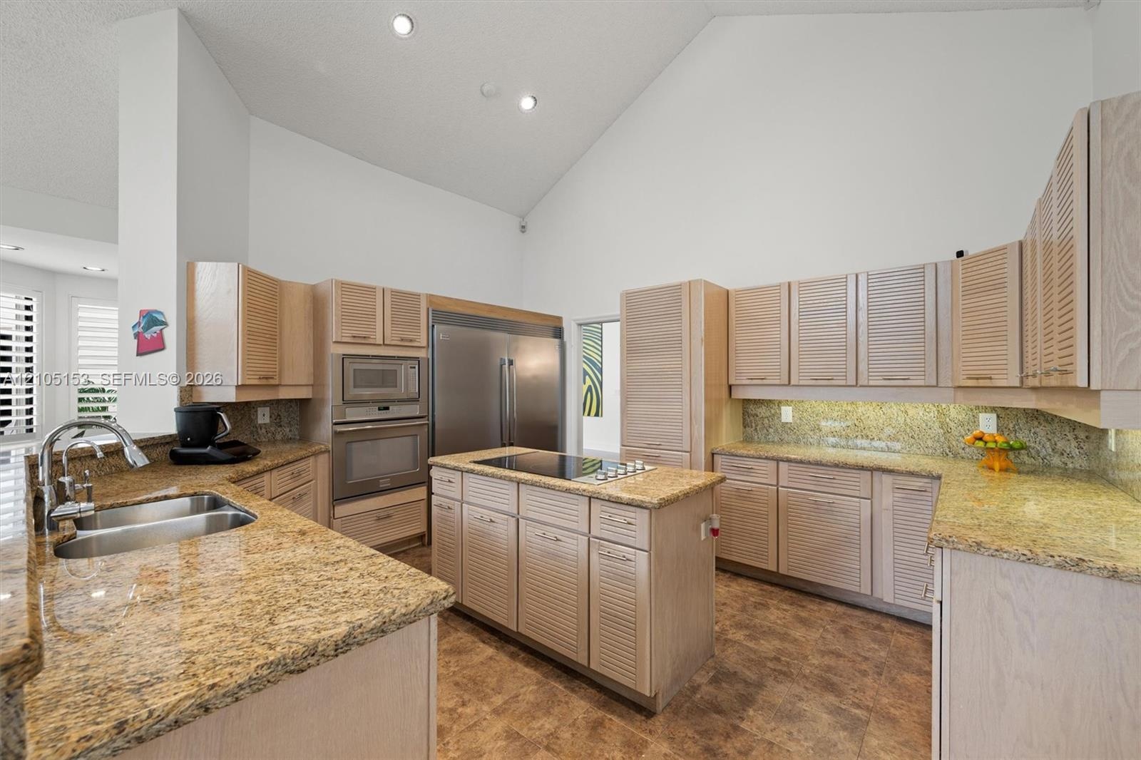 Kitchen featuring high vaulted ceilings, light wood-finish cabinetry, granite countertops, a stainless steel refrigerator, and a kitchen island with c
