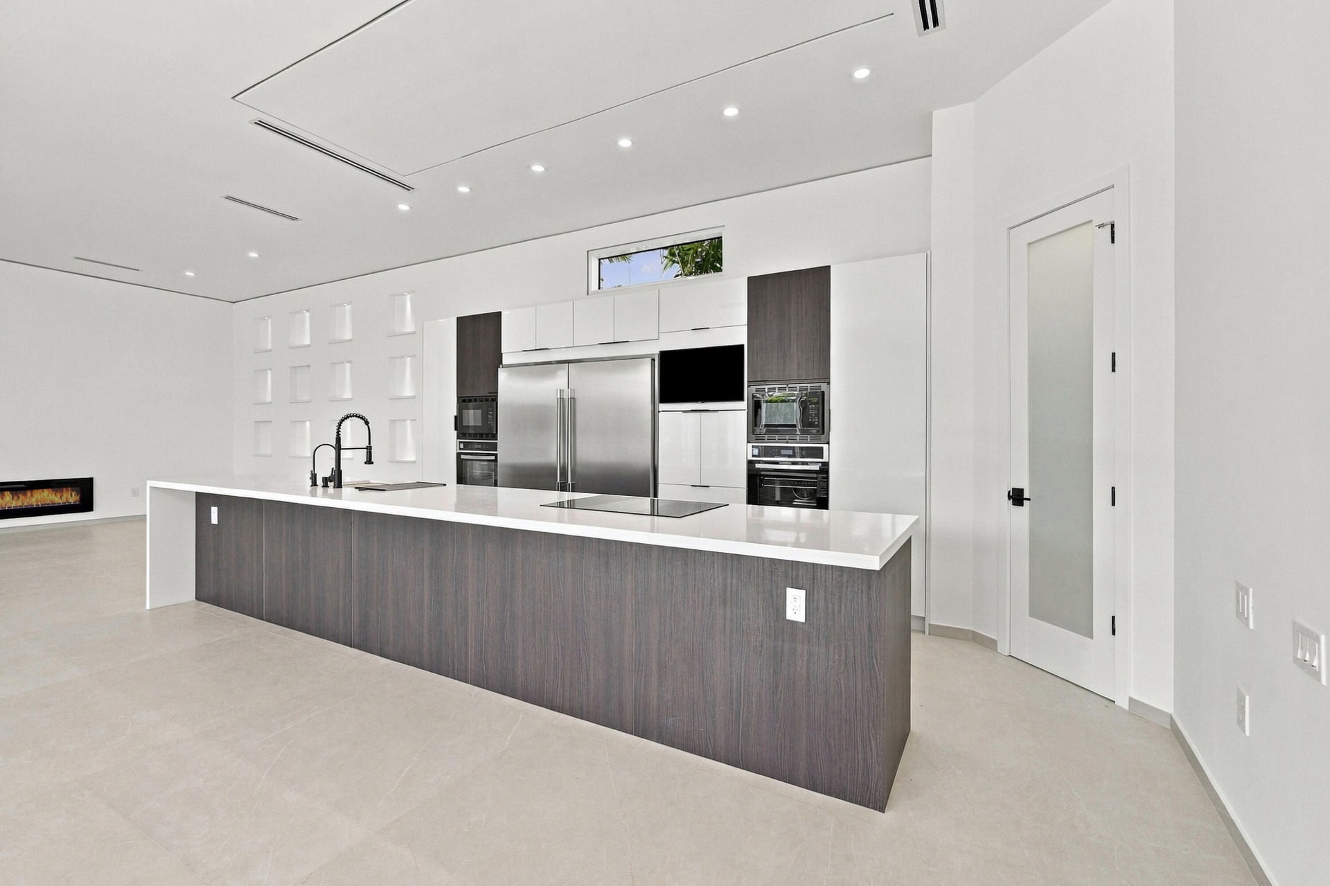 Contemporary kitchen featuring a large waterfall-edge island with dark wood-finish cabinetry, stainless steel appliances including a built-in refriger