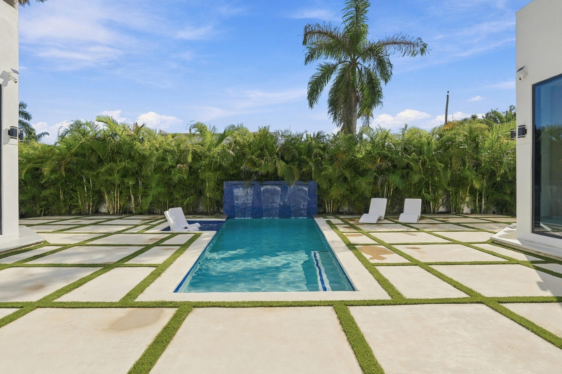 Rectangular swimming pool with a tile waterfall feature, surrounded by paver patio with artificial grass accents, and a lush privacy hedge