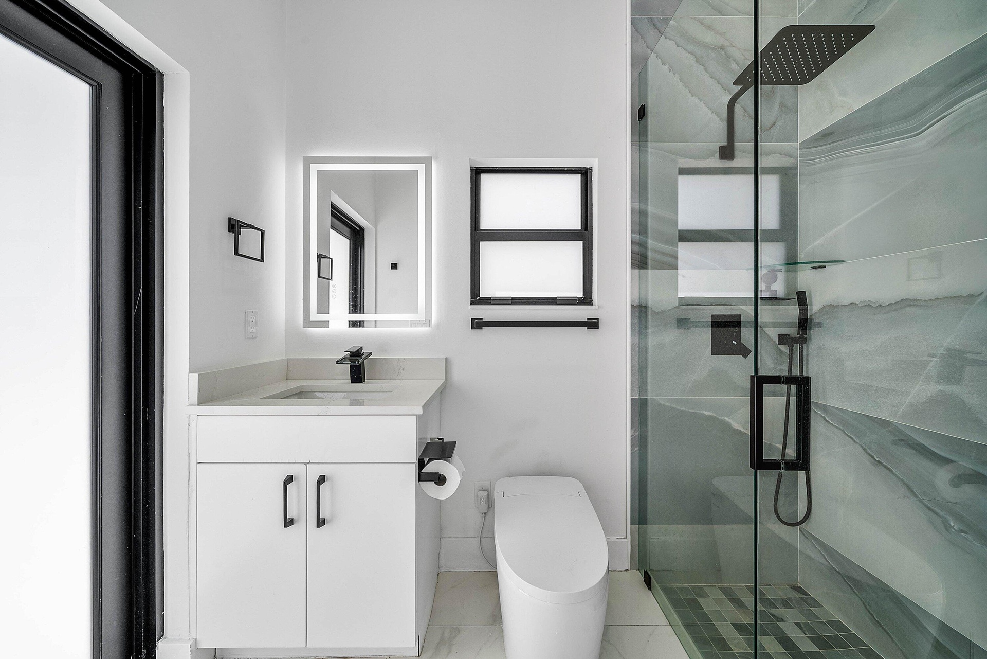 Frameless glass shower enclosure with a rainfall showerhead, white vanity with a light-colored countertop, and an illuminated mirror