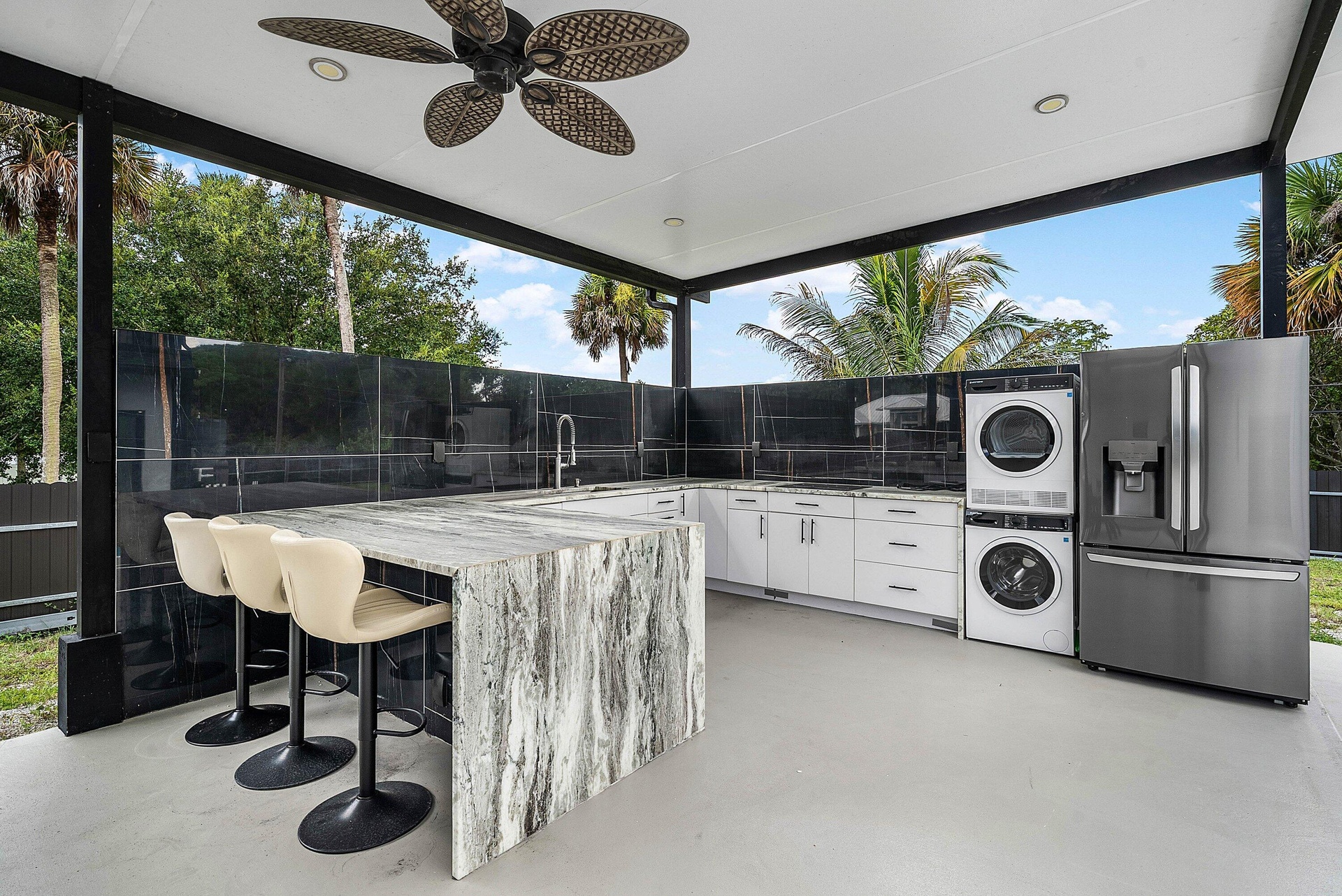 Covered outdoor kitchen featuring a large stone-finish island, white cabinetry, dark tile backsplash, a stacked washer and dryer, and a stainless stee