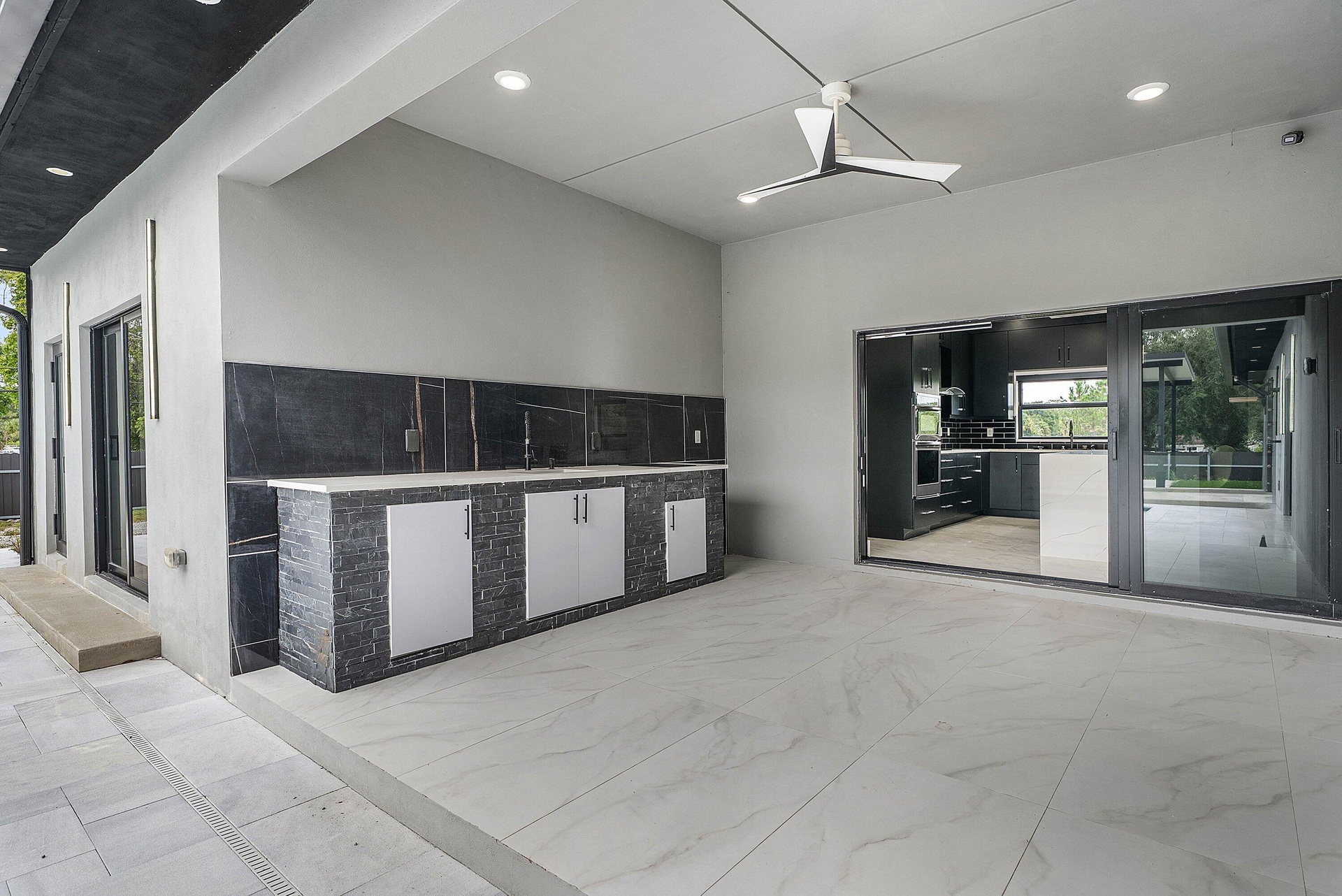 Covered outdoor living space featuring an integrated outdoor kitchen, contemporary ceiling fan, recessed lighting, and large format floor tiling