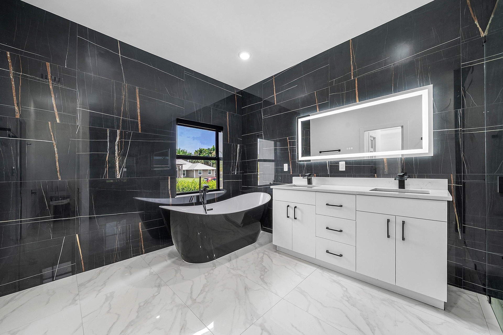Black-tiled walls with gold veining, a freestanding black soaking tub, a dual vanity with white cabinetry, a light-framed mirror, and large format whi