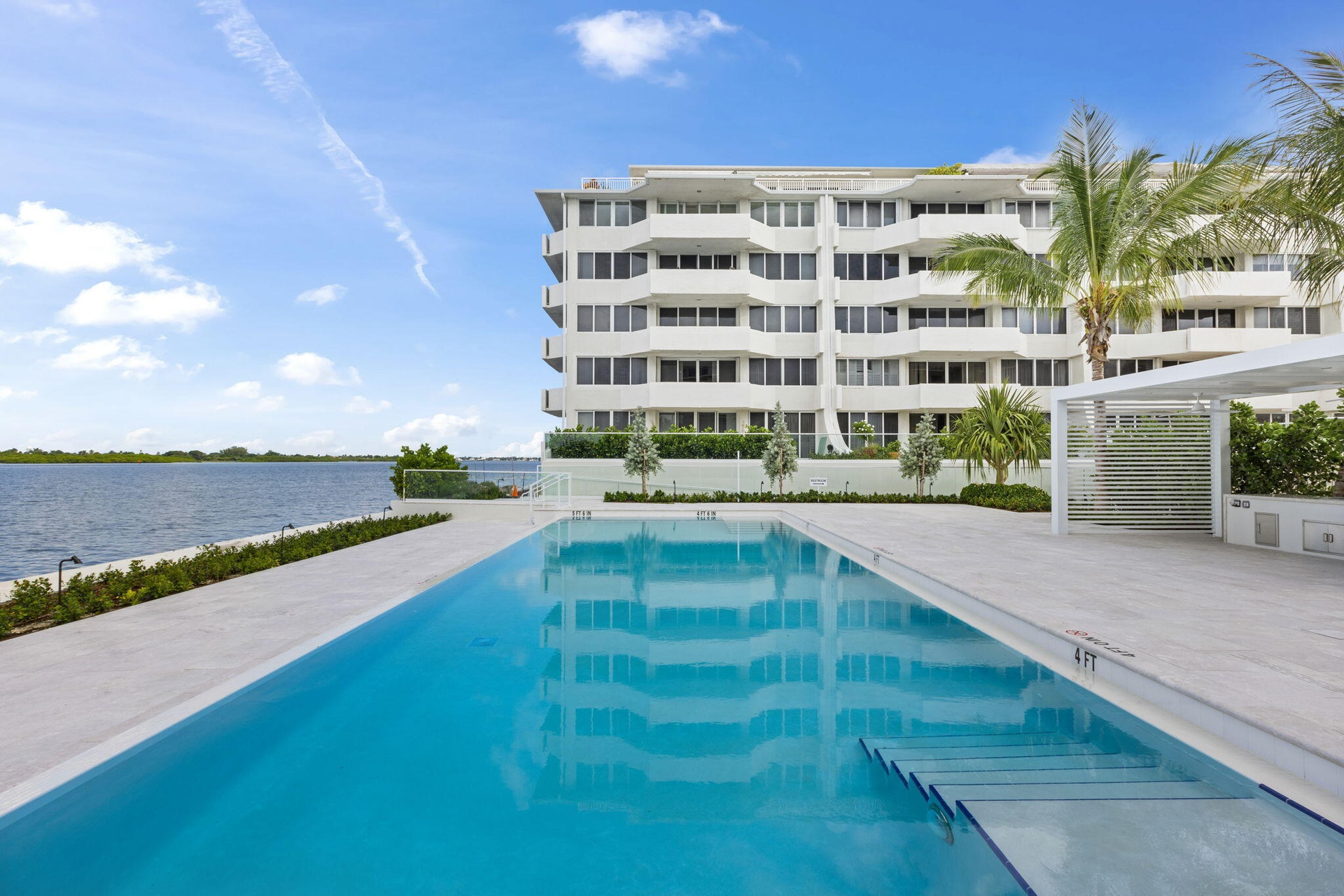 Modern high-rise residential building with multiple balconies, expansive outdoor swimming pool with submerged steps, white hardscaping, and a contempo