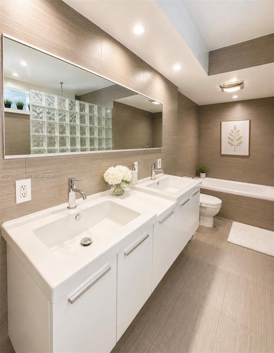 Dual vanity with integrated sinks and chrome fixtures, large rectangular mirror, recessed lighting, and a glass block shower wall