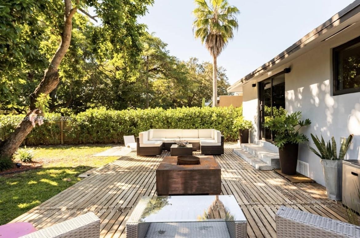 Expansive backyard featuring a wood-slat patio, a manicured lawn, lush perimeter hedging, and a white stucco exterior with dark-framed sliding glass d