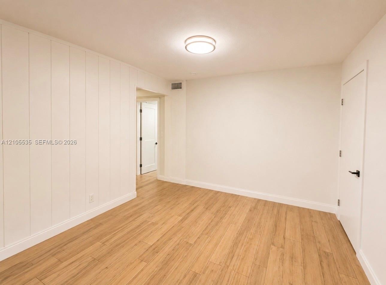 Wood-finish flooring, white paneled wall, flat panel interior door, ceiling-mounted light fixture, white baseboards