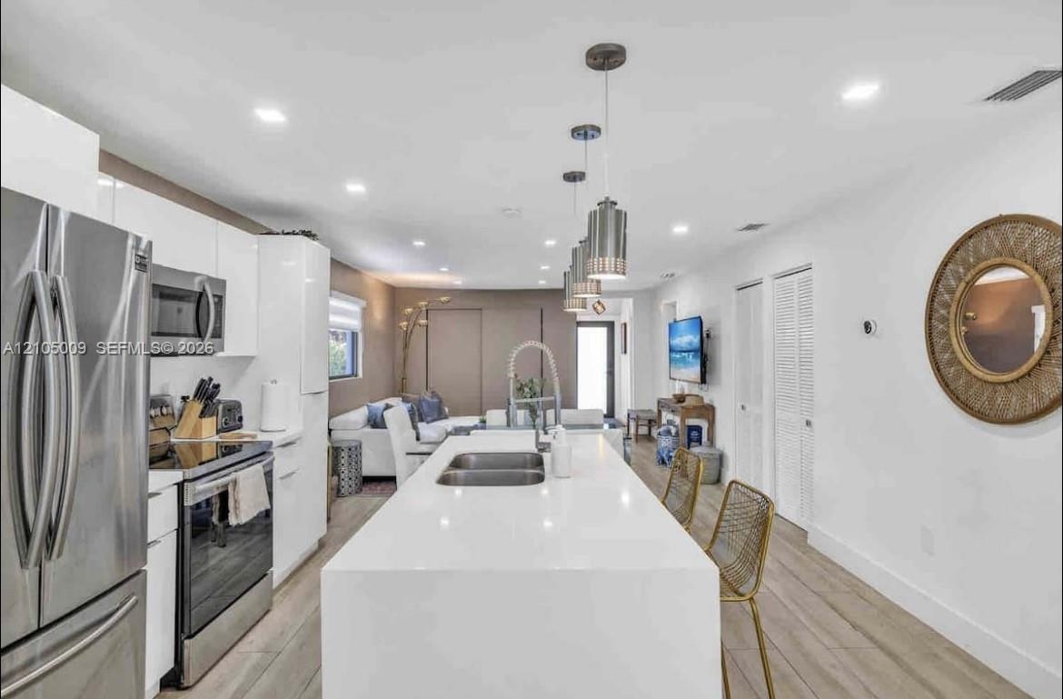 Modern kitchen featuring a waterfall island with an undermount double sink, stainless steel appliances, white flat-panel cabinetry, and wood-finish fl