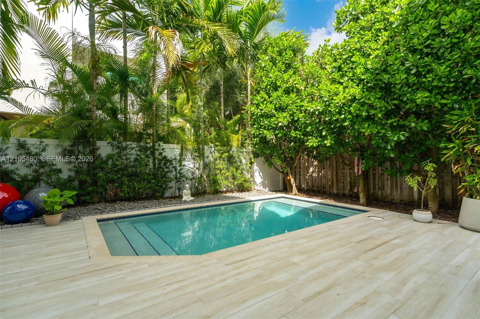 Private backyard oasis featuring a swimming pool with integrated steps, surrounded by light-toned plank decking, lush tropical landscaping, and a priv