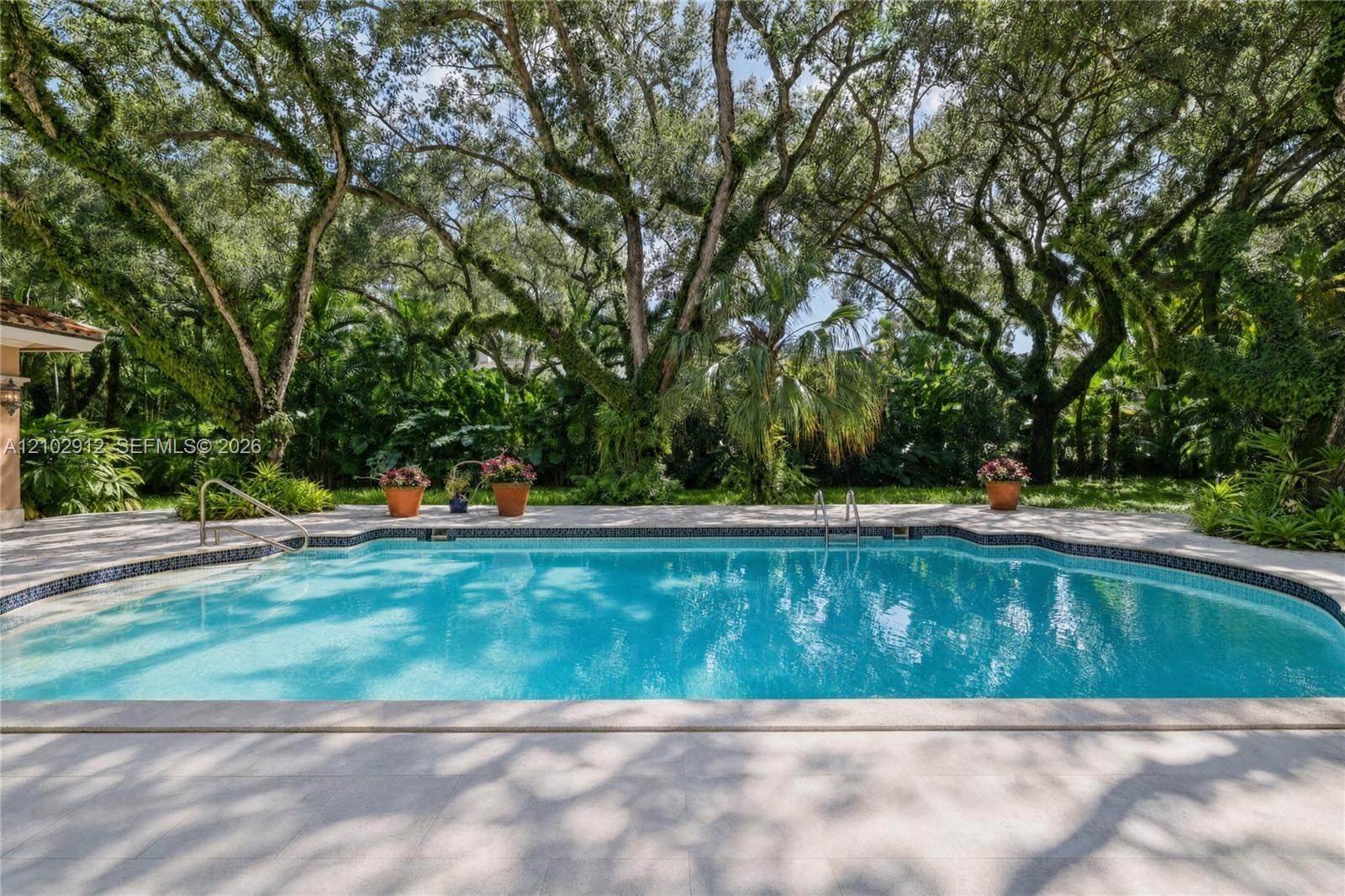 Freeform swimming pool with a tiled perimeter, integrated steps, and stainless steel handrails, surrounded by extensive hardscaping and mature trees