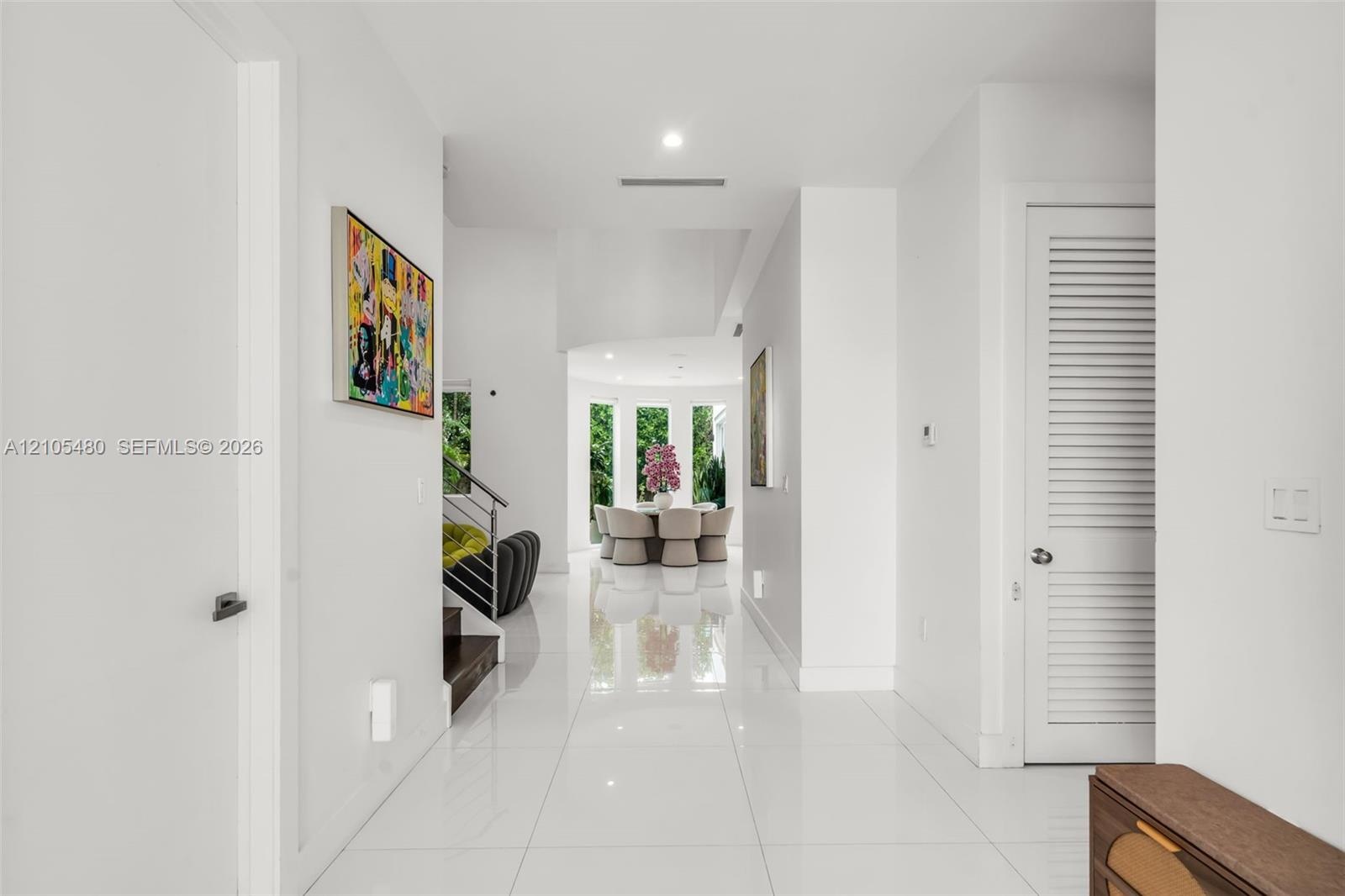 Expansive hallway featuring polished tile flooring, recessed lighting, white walls, a built-in staircase with a metal handrail, and a louvered door