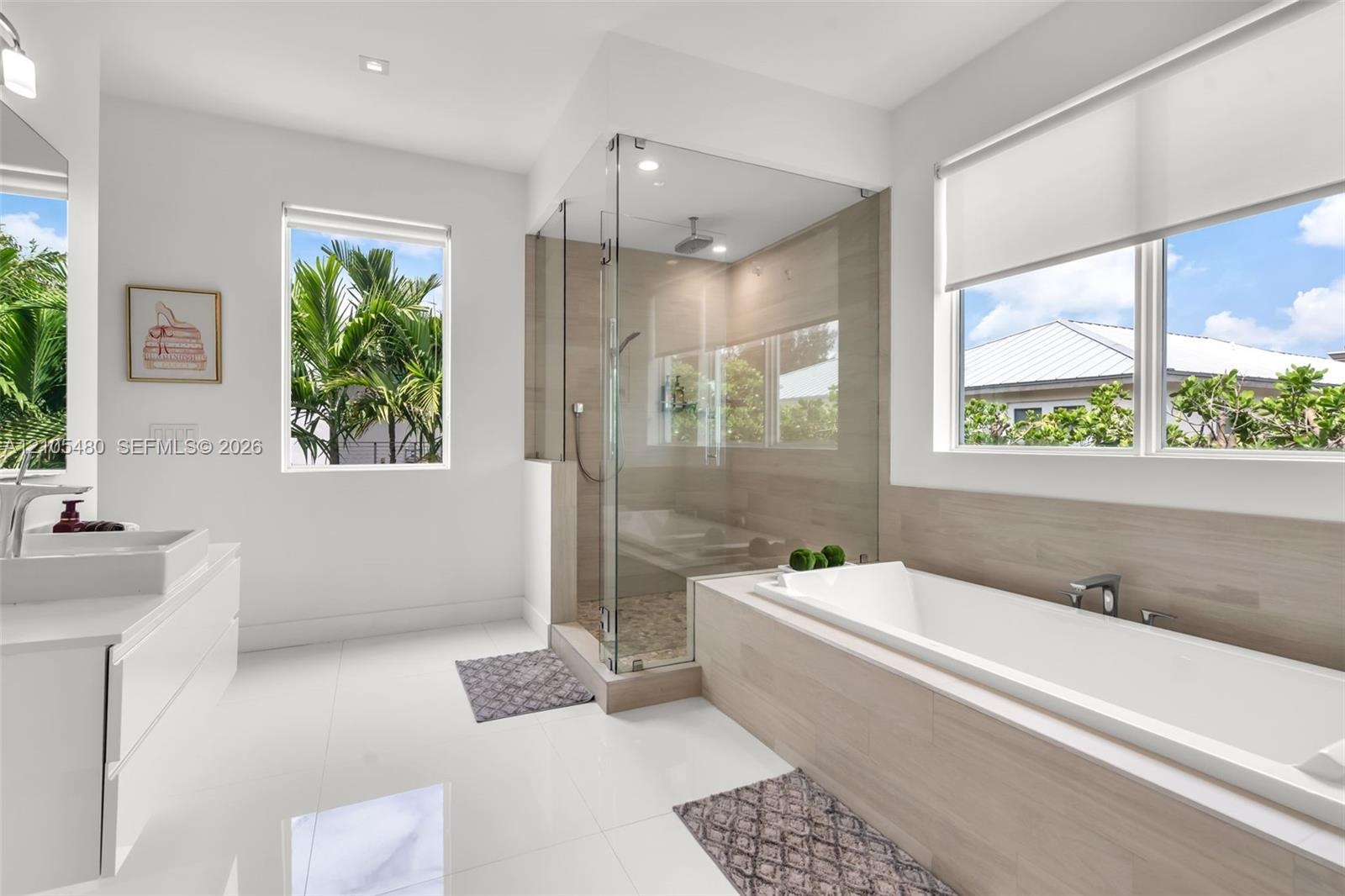 Spacious bathroom featuring white floor tile, a soaking tub with wood-finish surround, a glass-enclosed shower, dual windows, and a floating vanity wi
