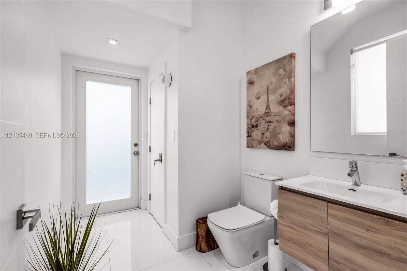 White tile flooring, contemporary vanity with wood-finish cabinetry, rectangular under-mount sink, chrome faucet, and a wall-mounted mirror with integ