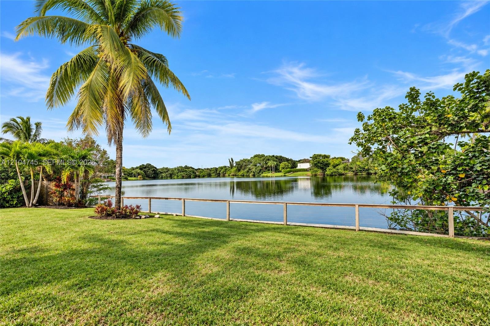 Expansive lawn with a wooden fence bordering a lake, mature palm trees, and established landscaping
