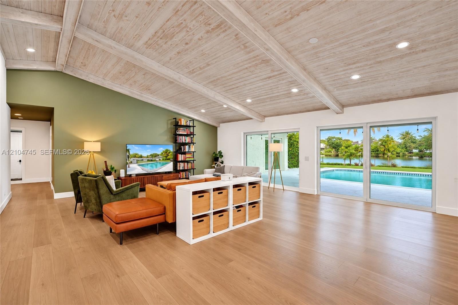 Vaulted ceiling with exposed wood beams, recessed lighting, wood-finish flooring, and expansive sliding glass doors opening to a private pool and wate