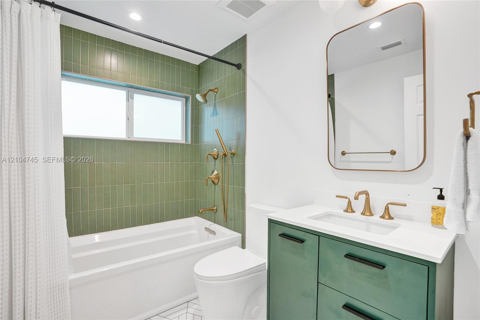 Bathtub with green vertical tile surround, gold-finish shower and bath fixtures, white vanity with green cabinetry, white countertop, and gold-finish 