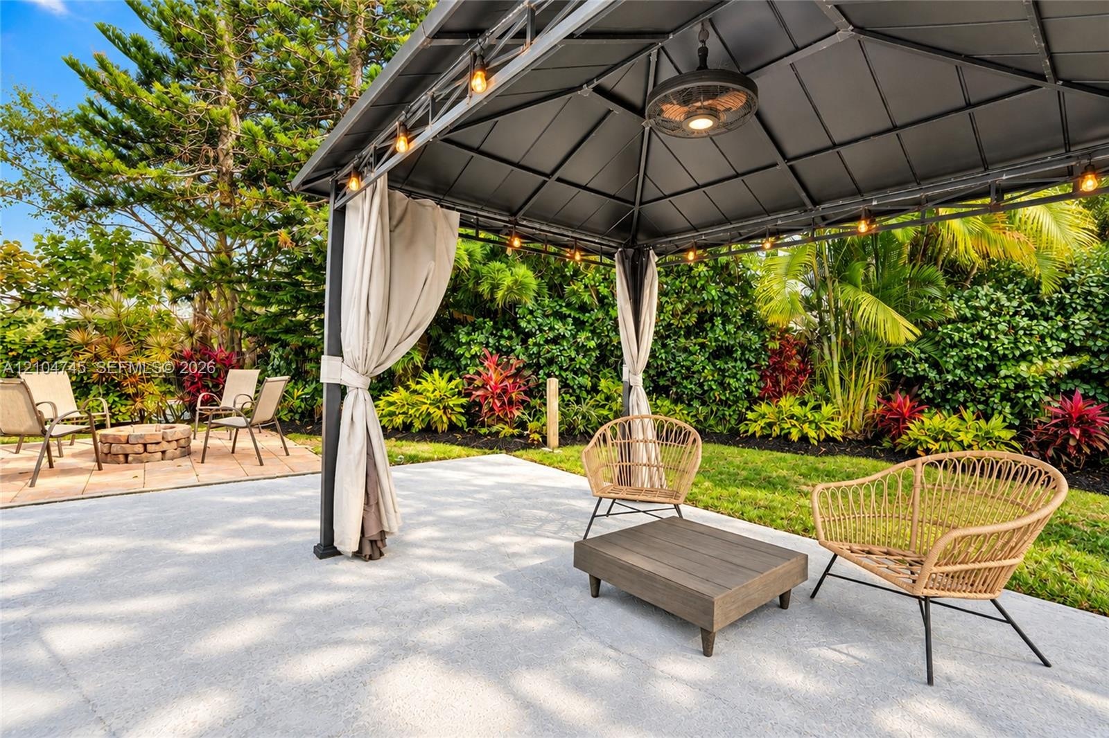 Spacious patio featuring a gazebo with integrated lighting and ceiling fan, a separate paver-stone area with a fire pit, and lush, mature landscaping