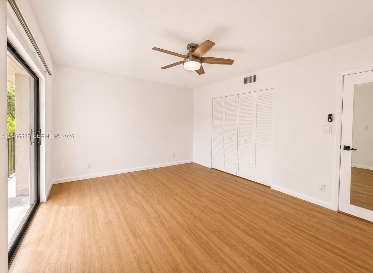 Wood-finish flooring throughout, featuring a ceiling fan with integrated lighting, sliding glass door with balcony, louvered bi-fold closet doors, and