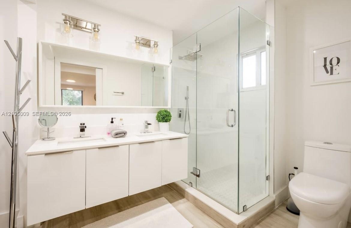 Dual sink vanity with white cabinetry and integrated sinks, large mirrored wall, glass-enclosed shower with tiled walls and floor, and wood-finish flo