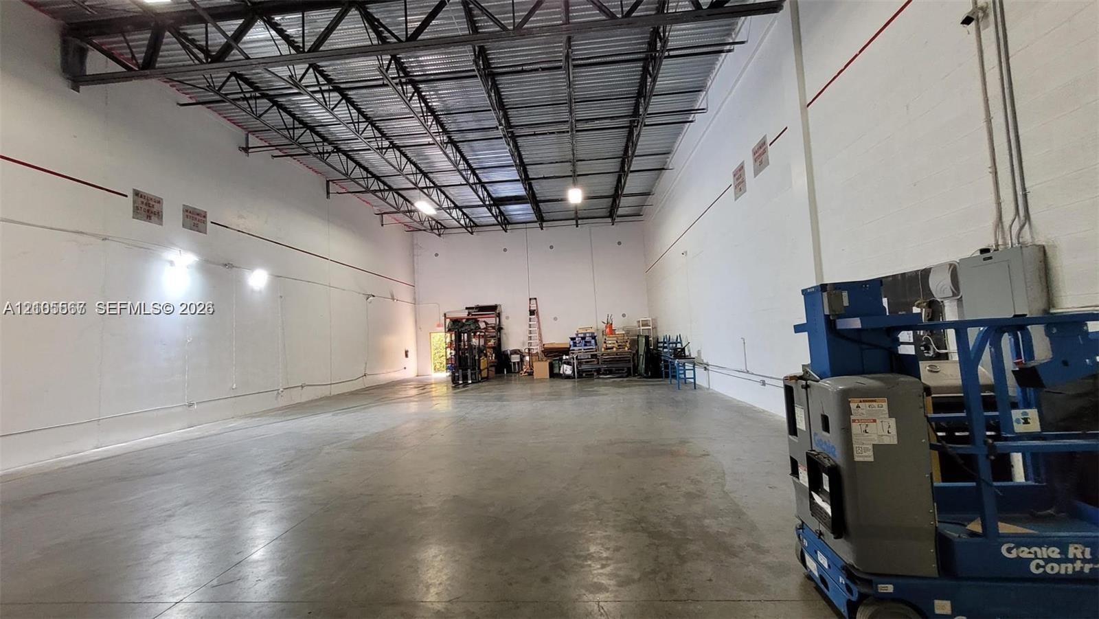 Spacious interior featuring a concrete floor, high exposed metal beam ceiling, white walls, and industrial lighting fixtures