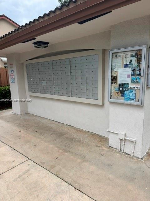 Mailbox station featuring multiple lockable compartments, exterior lighting, a tiled roof overhang, and a community bulletin board