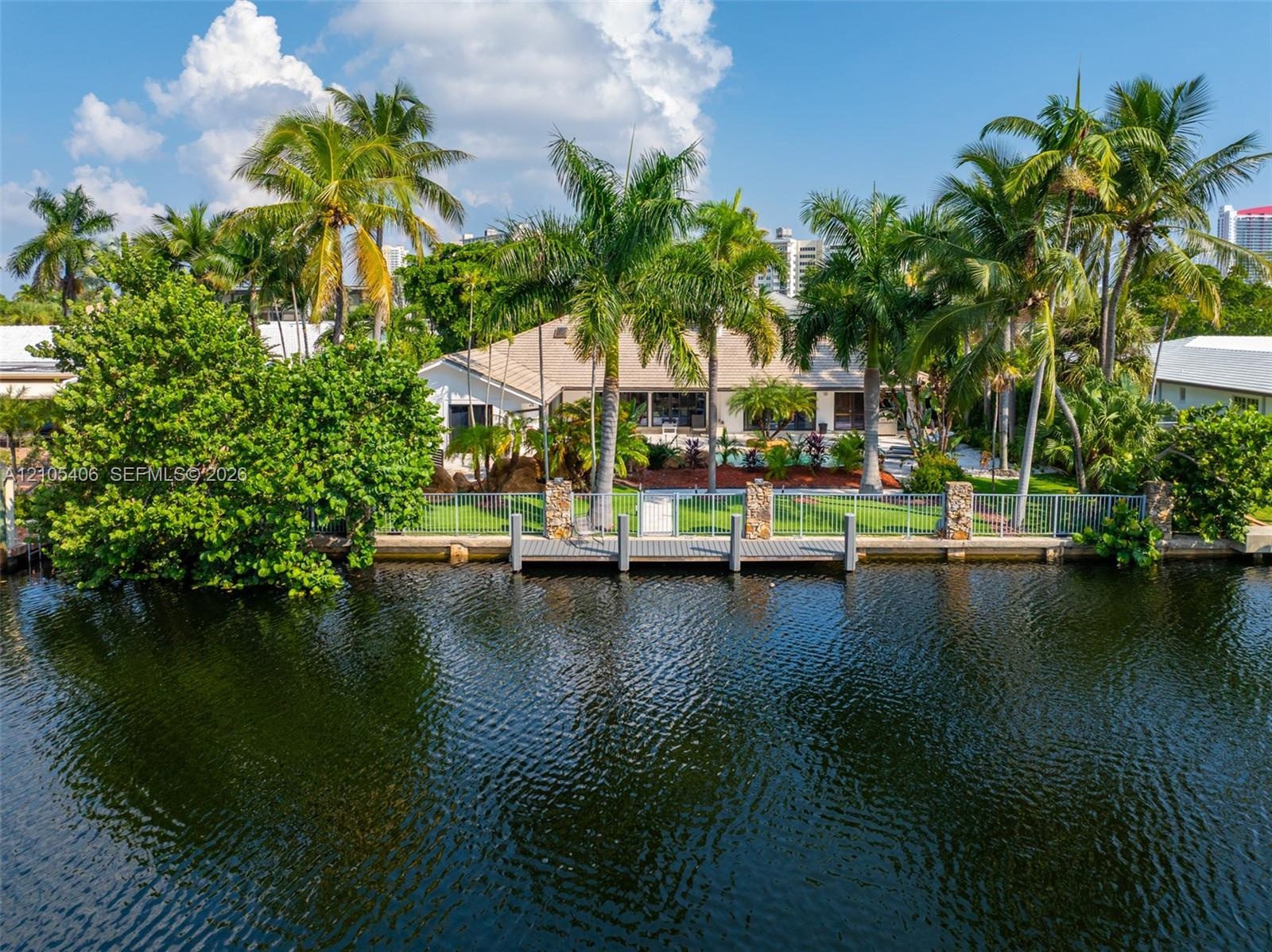 Waterfront property featuring a private dock with pilings, a manicured lawn, mature palm trees, and established landscaping