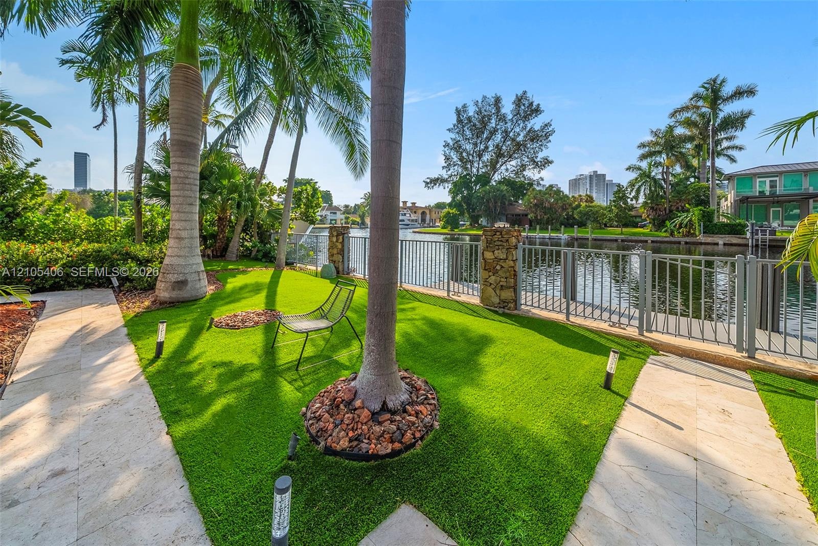 Waterfront property featuring a metal fence with stone pillars, a stone pathway, artificial turf, and mature palm trees