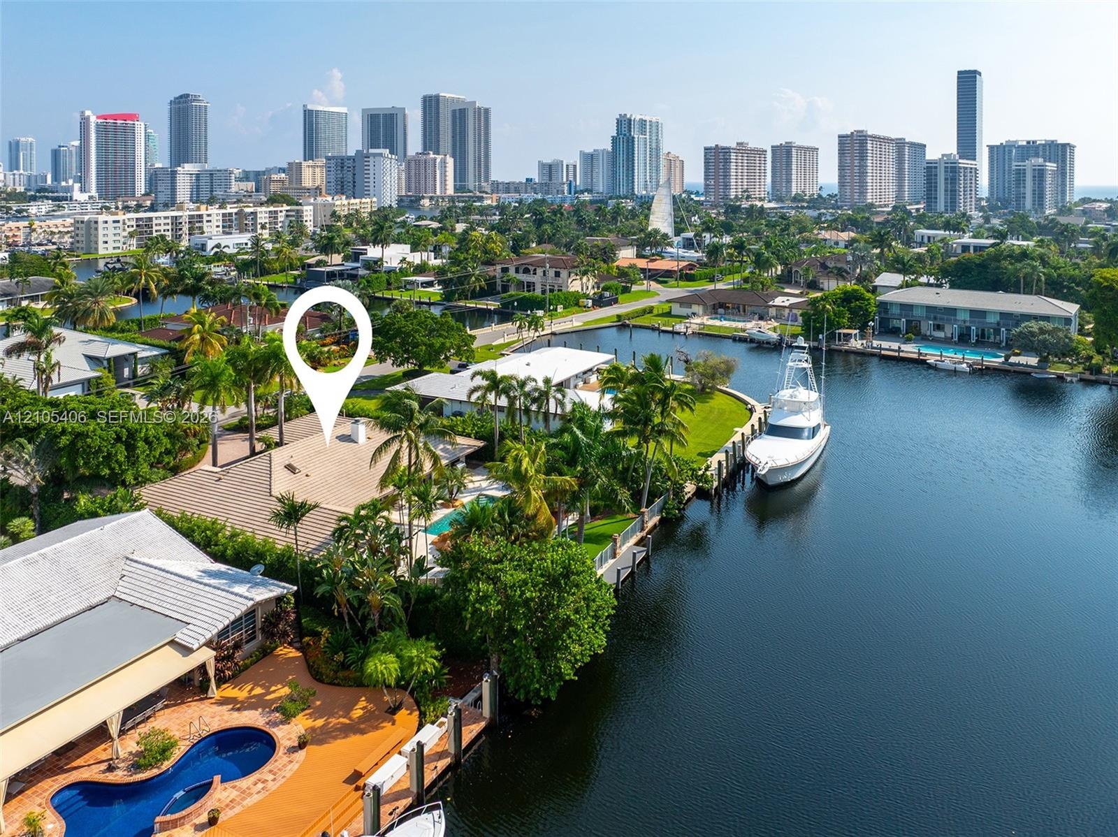 Waterfront property featuring a private boat dock, lush landscaping with mature palm trees, a swimming pool, and a tiled roof, with a city skyline in 