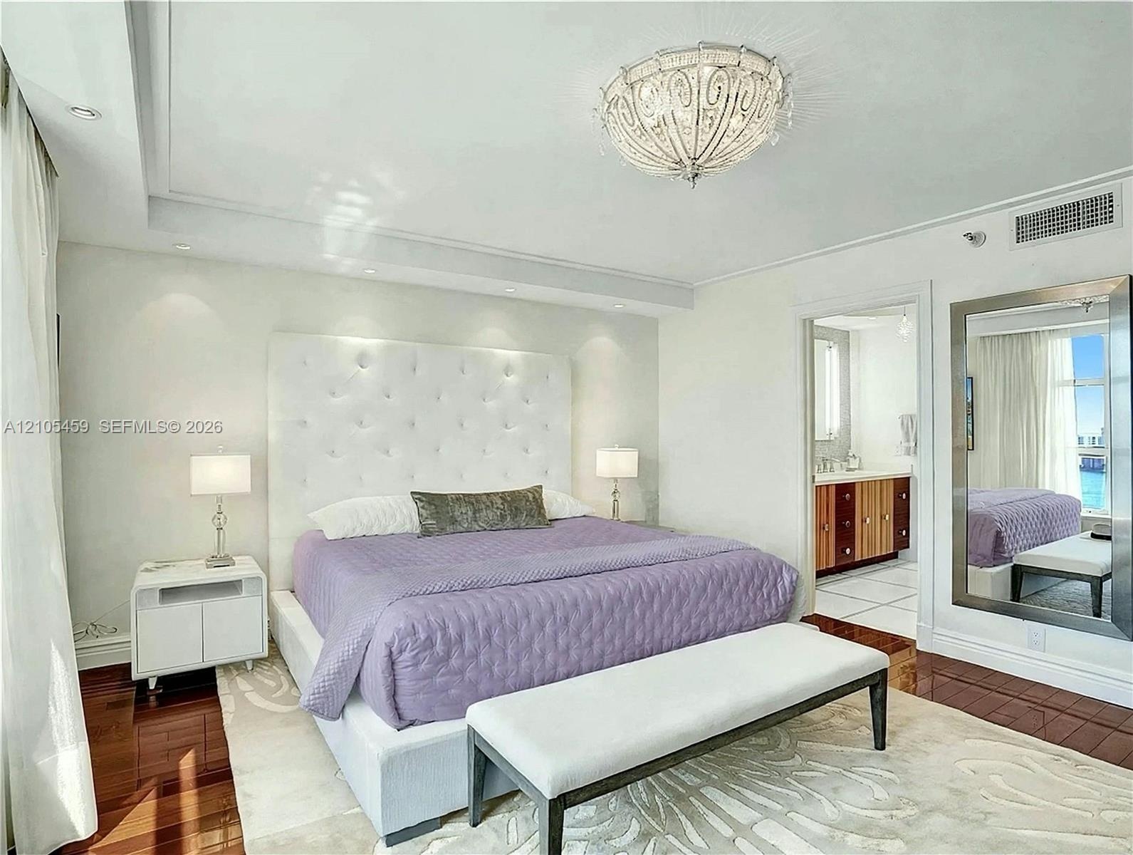 Tray ceiling with recessed lighting, ornate ceiling fixture, wood-finish flooring, tufted headboard, and an en-suite bathroom with a wood-finish vanit