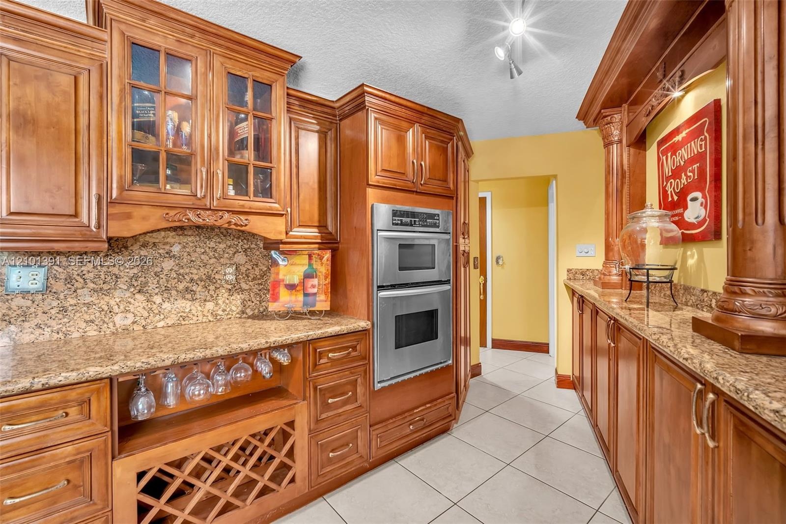 Custom wood cabinetry with glass-front accents, stainless steel double wall ovens, a built-in wine rack, and a stone-finish backsplash with countertop