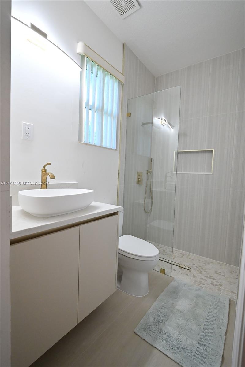 Floating vanity with a vessel sink and gold-tone faucet, glass-enclosed shower with a tiled wall and pebble floor, and wood-finish flooring