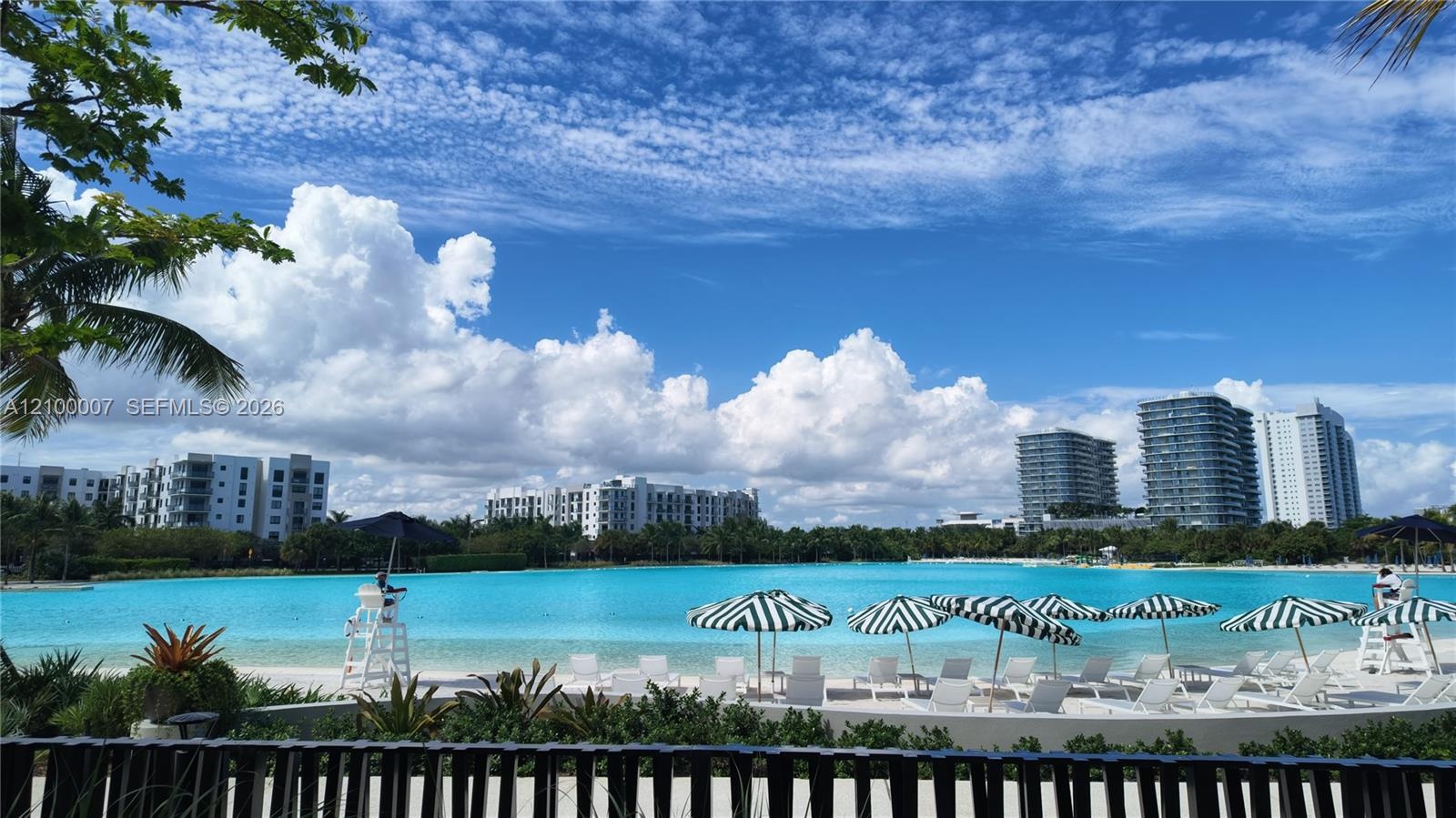 Expansive water feature with white sand shore, multiple striped umbrellas, and mature landscaping