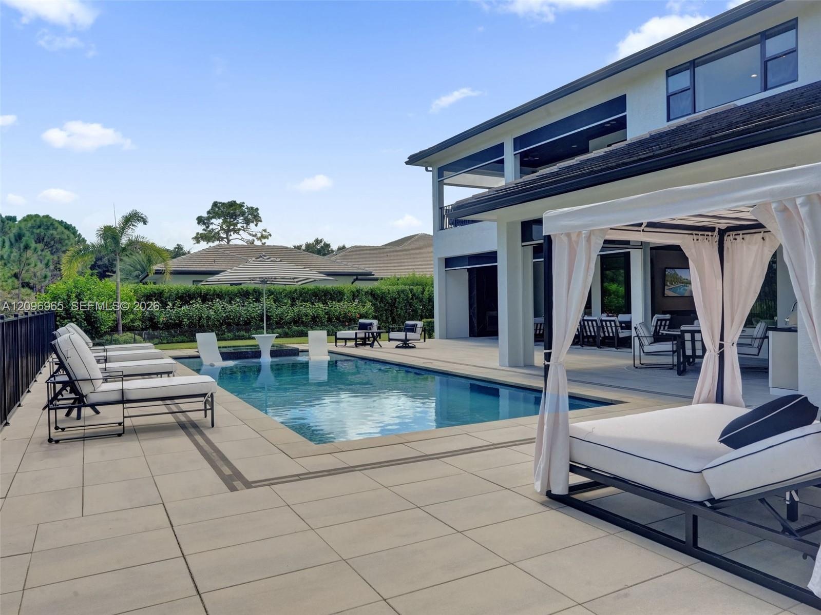 Outdoor patio with large-format tile flooring, rectangular in-ground pool, metal perimeter fencing, and mature pine trees