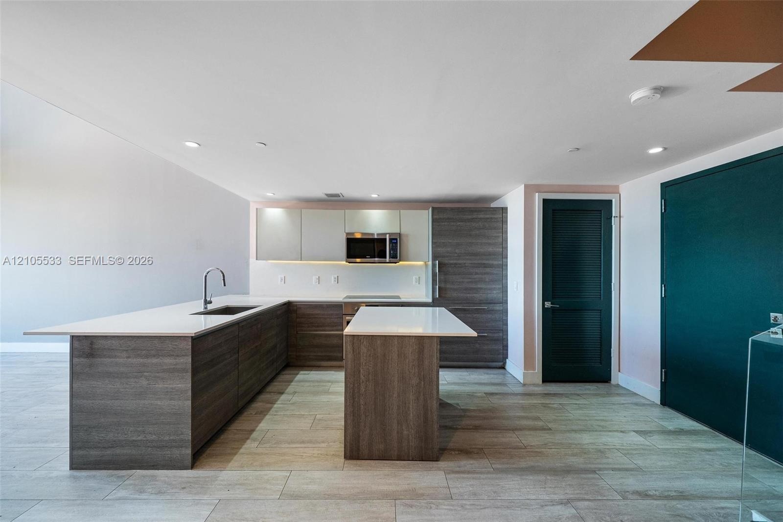 Kitchen featuring wood-finish cabinetry, an island with a light-toned countertop, undermount sink, built-in microwave, and tile flooring