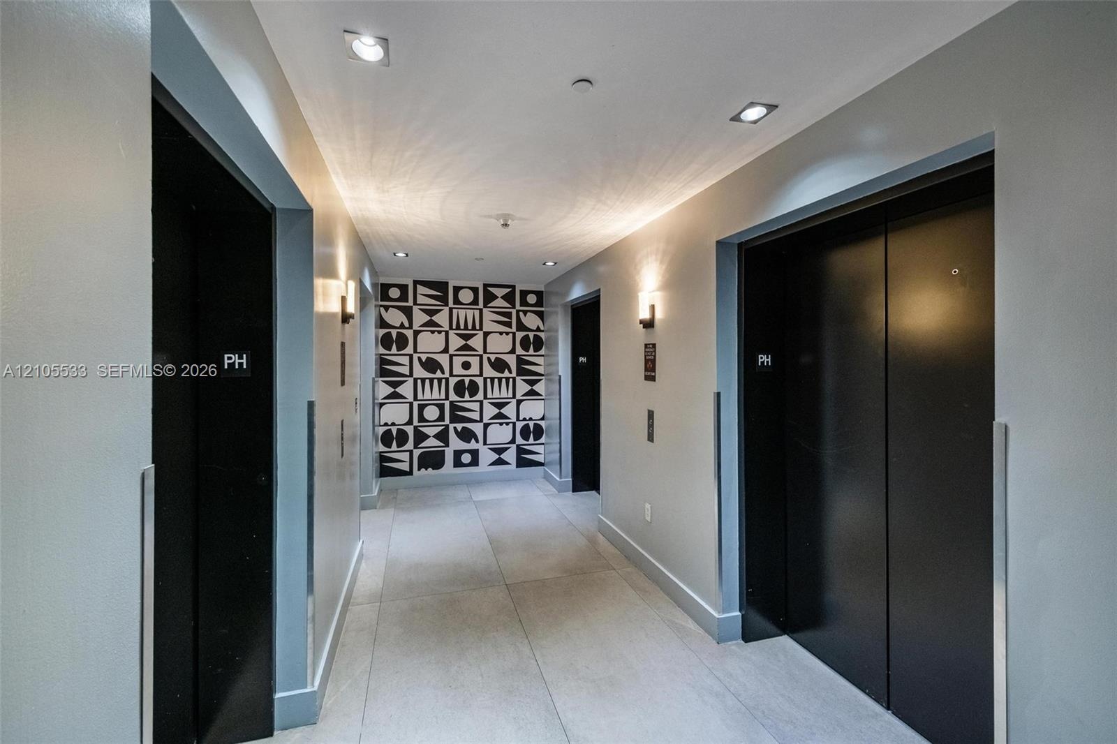 Elevator lobby featuring two matte black elevator doors, large-format tile flooring, recessed ceiling lighting, and a geometric-patterned accent wall