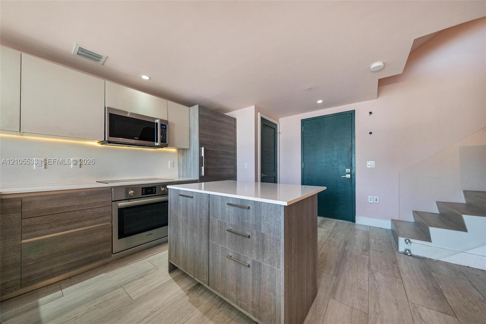 Kitchen featuring flat-panel cabinetry, a central island with a solid-surface countertop, stainless steel appliances, wood-finish flooring, and a glas