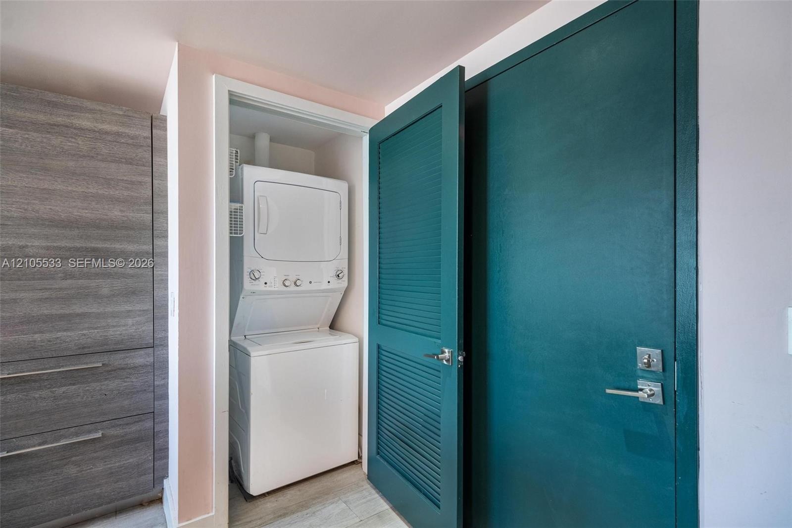 Stacked laundry unit housed in a dedicated closet, featuring a teal louvered door, wood-finish built-in cabinetry, and wood-finish flooring