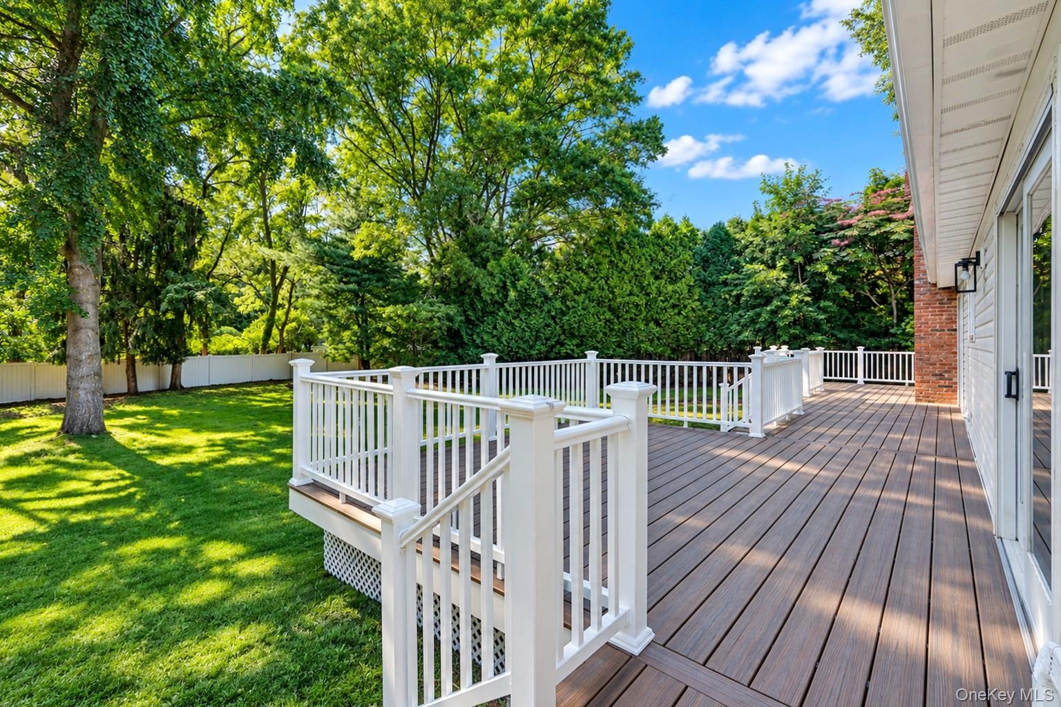 Raised composite deck with white railings, expansive grass lawn, mature trees, and white perimeter fencing